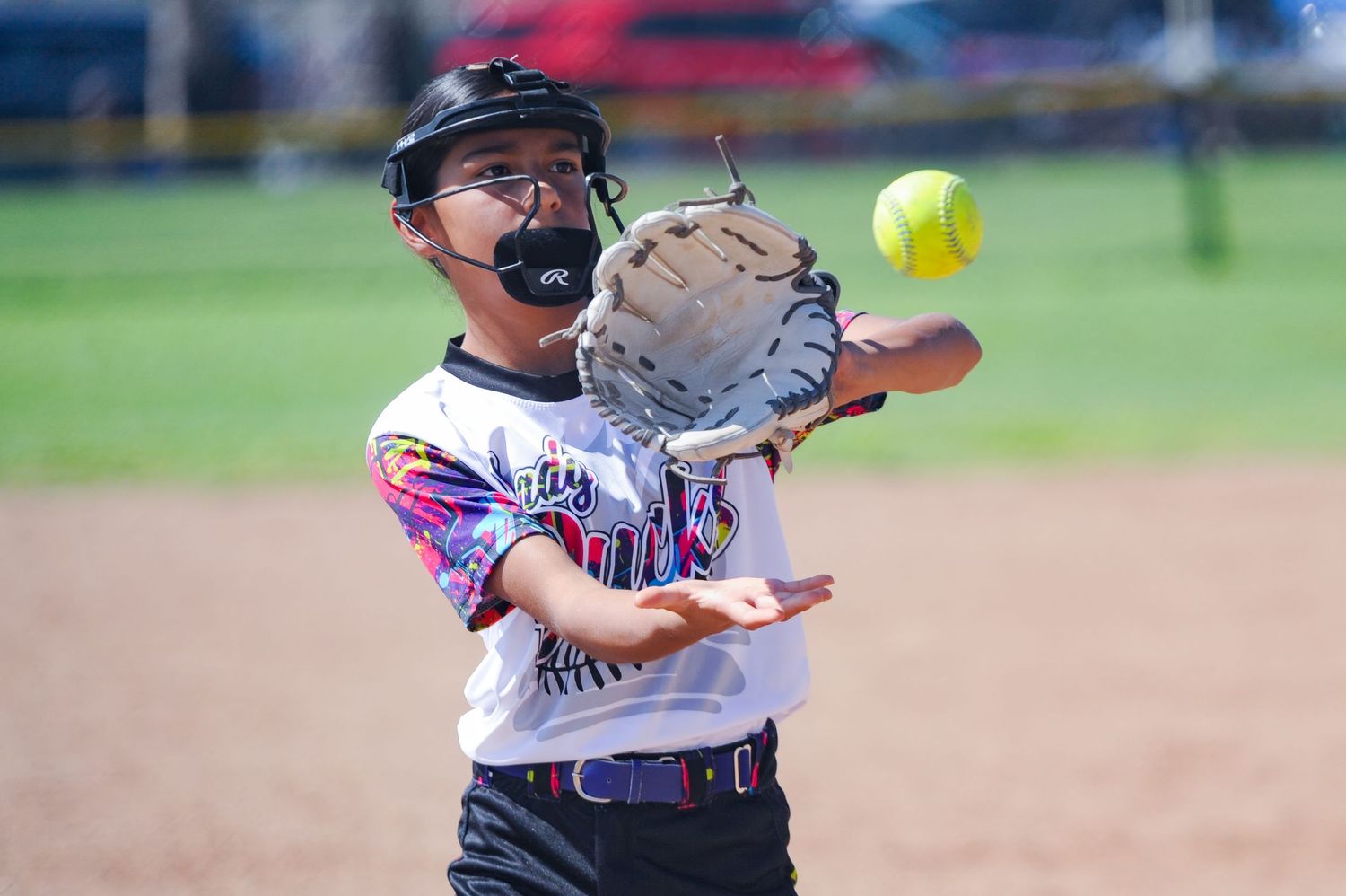 Young softball player in colorful jersey prepares to catch an incoming yellow softball.