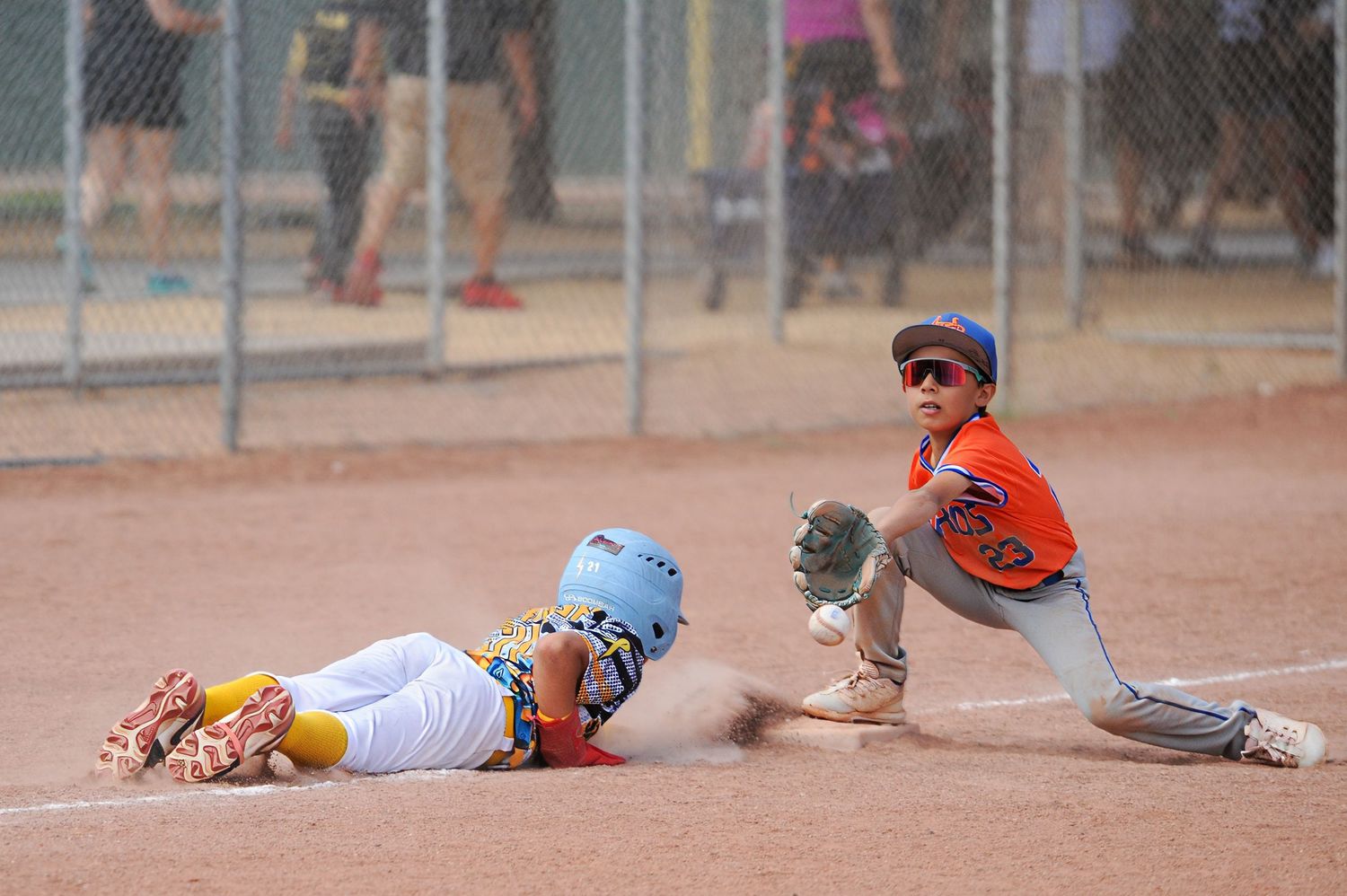 Youth baseball player slides into base while fielder in orange uniform attempts a tag play.