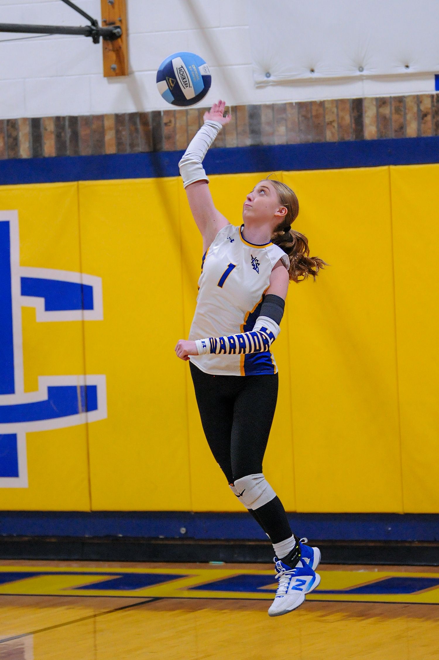 Female volleyball player in Warriors uniform jumping to serve a blue and white ball in a yellow gymnasium.