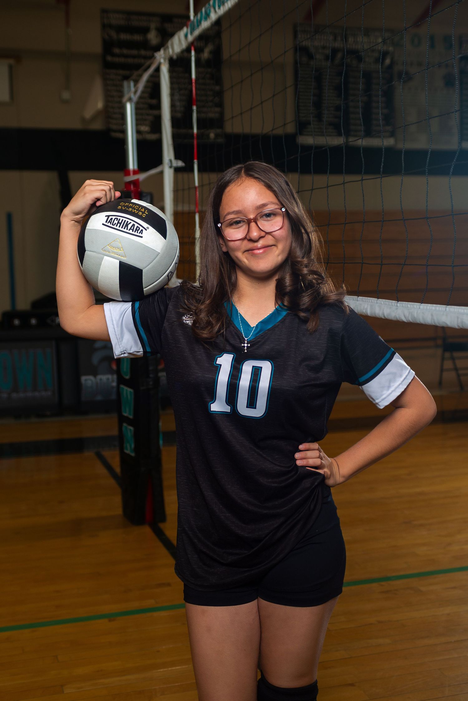 Young female volleyball player #10 poses with ball beside net in black jersey in gymnasium.