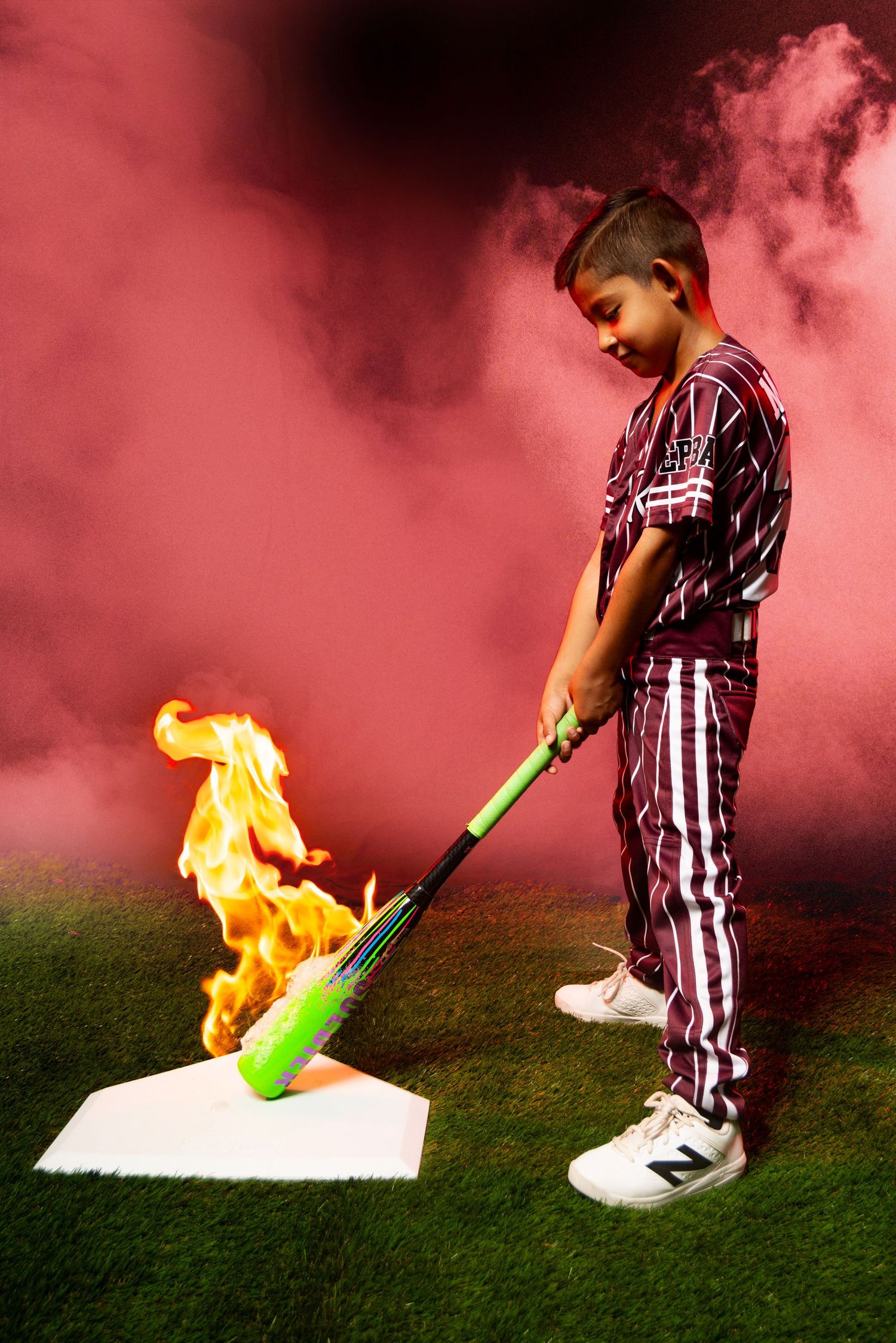 Young baseball player in maroon pinstripe uniform swings bat at flaming home plate with pink smoke background.
