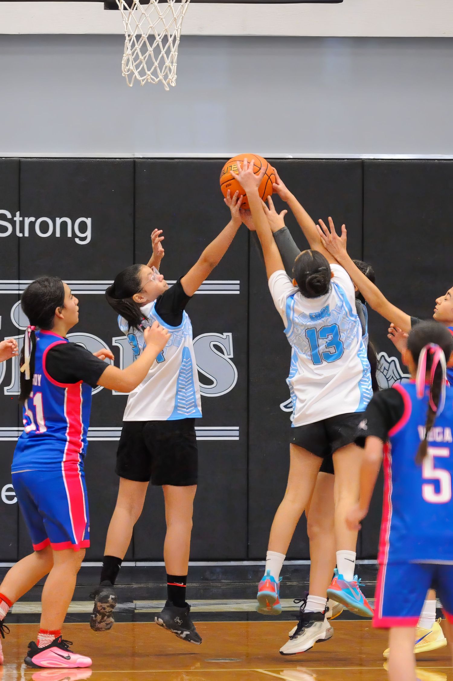 Girls basketball players compete for a rebound during an indoor high school game, with players in blue and white uniforms.
