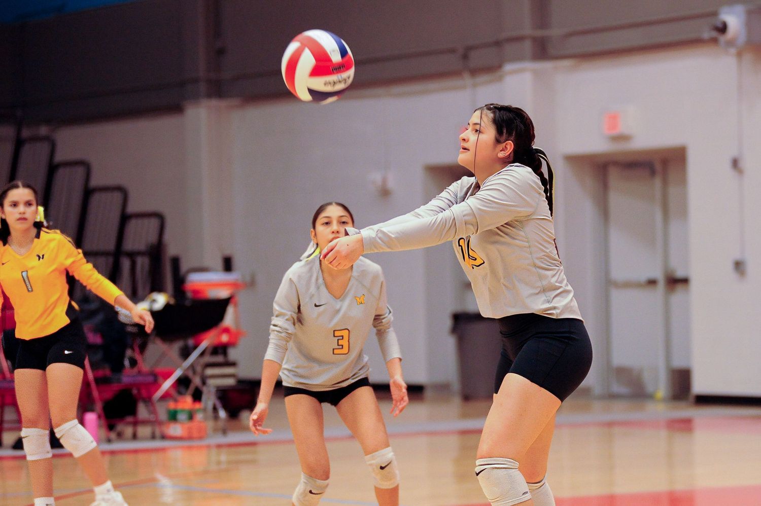 Female volleyball player performs a forearm pass during an indoor match, with teammates visible in the background.
