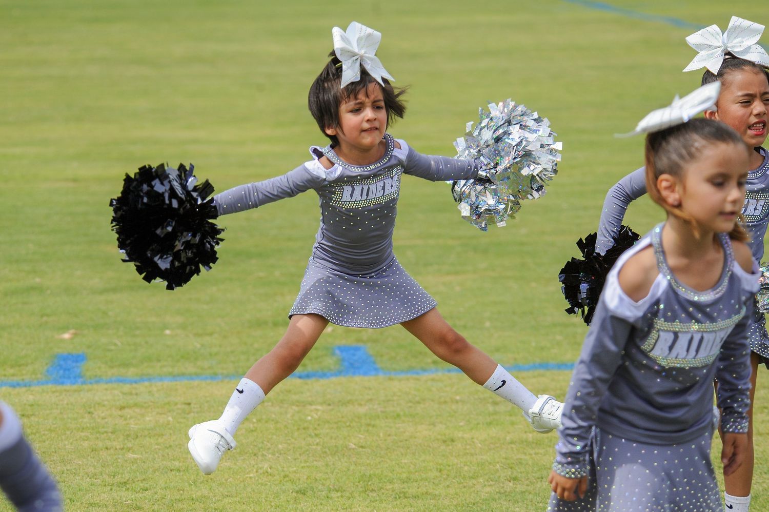 Young girl cheerleader in gray Raiders uniform jumps mid-air holding black and silver pom-poms on green field.