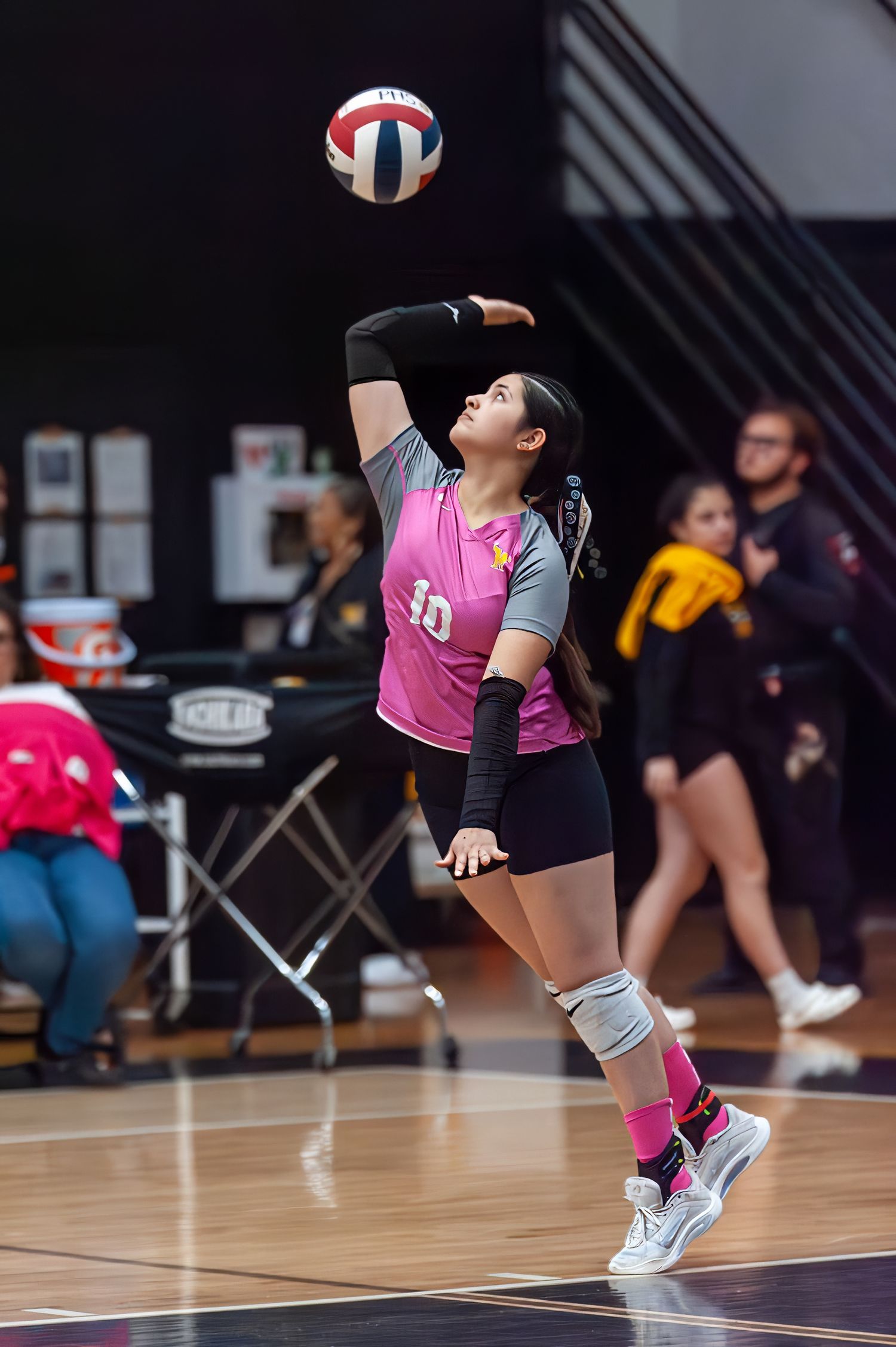 Female volleyball player #10 in pink jersey serves ball during indoor match, wearing knee pads and athletic shoes.
