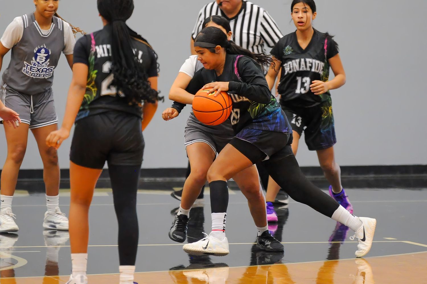 Girls youth basketball players compete in a game, with one player dribbling while defended by opponents in black uniforms.
