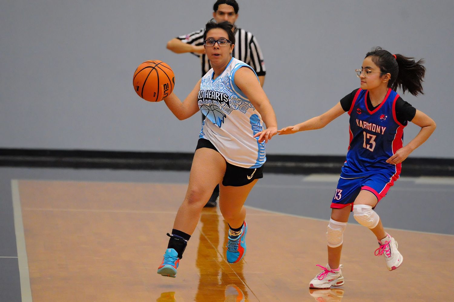 Two female basketball players compete on indoor court, one dribbling in white uniform, other defending in red number 13 jersey.