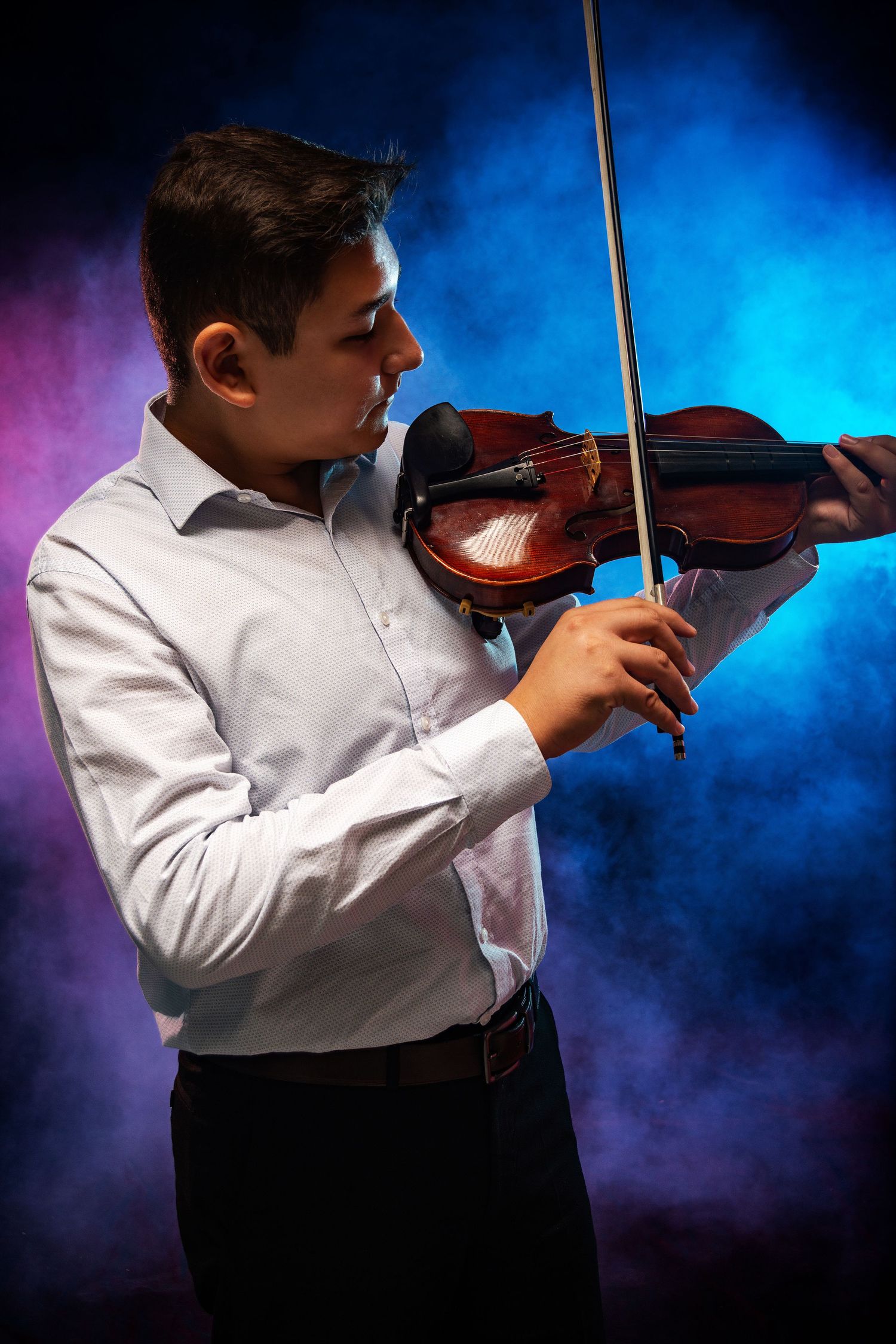 Young male violinist in white shirt playing violin against dramatic blue and pink smoky background.