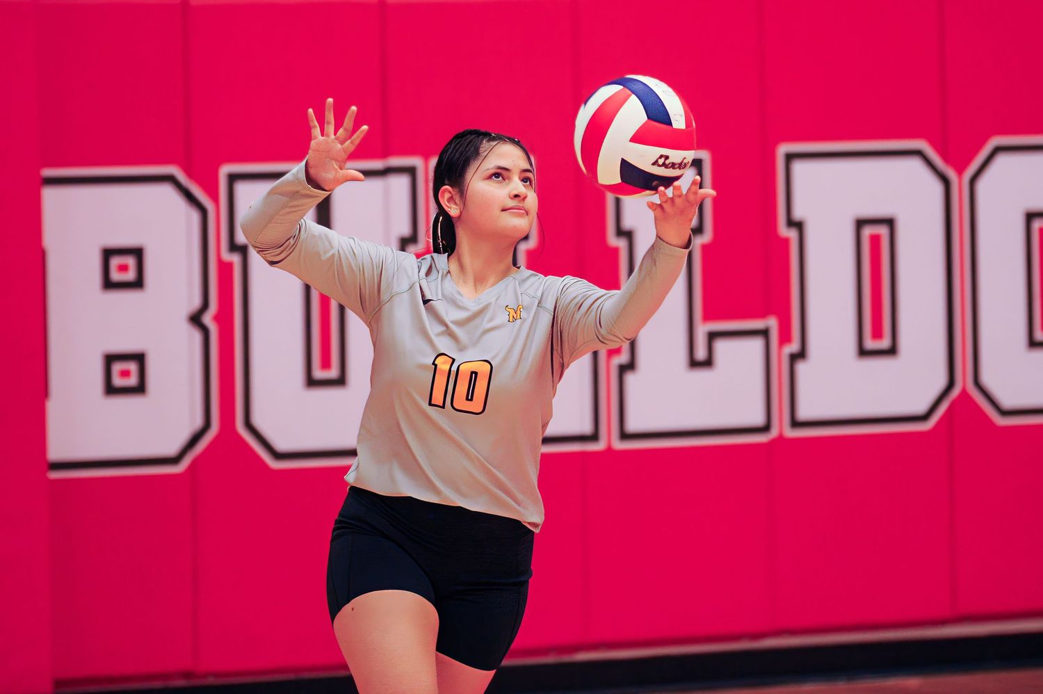 Female volleyball player wearing number 10 jersey prepares to serve ball during indoor match in front of Bulldogs sign.