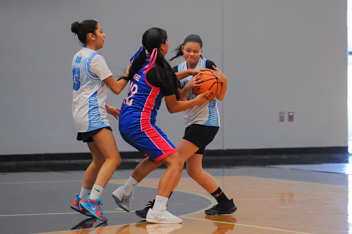 Girls youth basketball players competing in a gym, with two defenders guarding a player in a red uniform holding the ball.