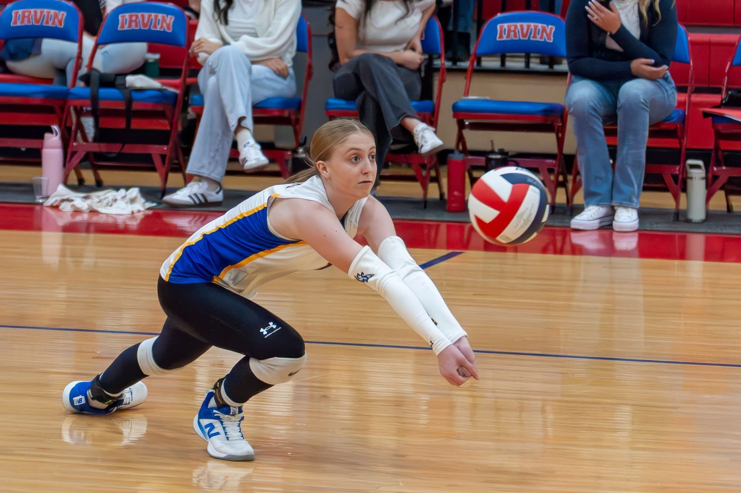 Female volleyball player in blue uniform performs a dig, lunging low to pass the ball during an indoor match.