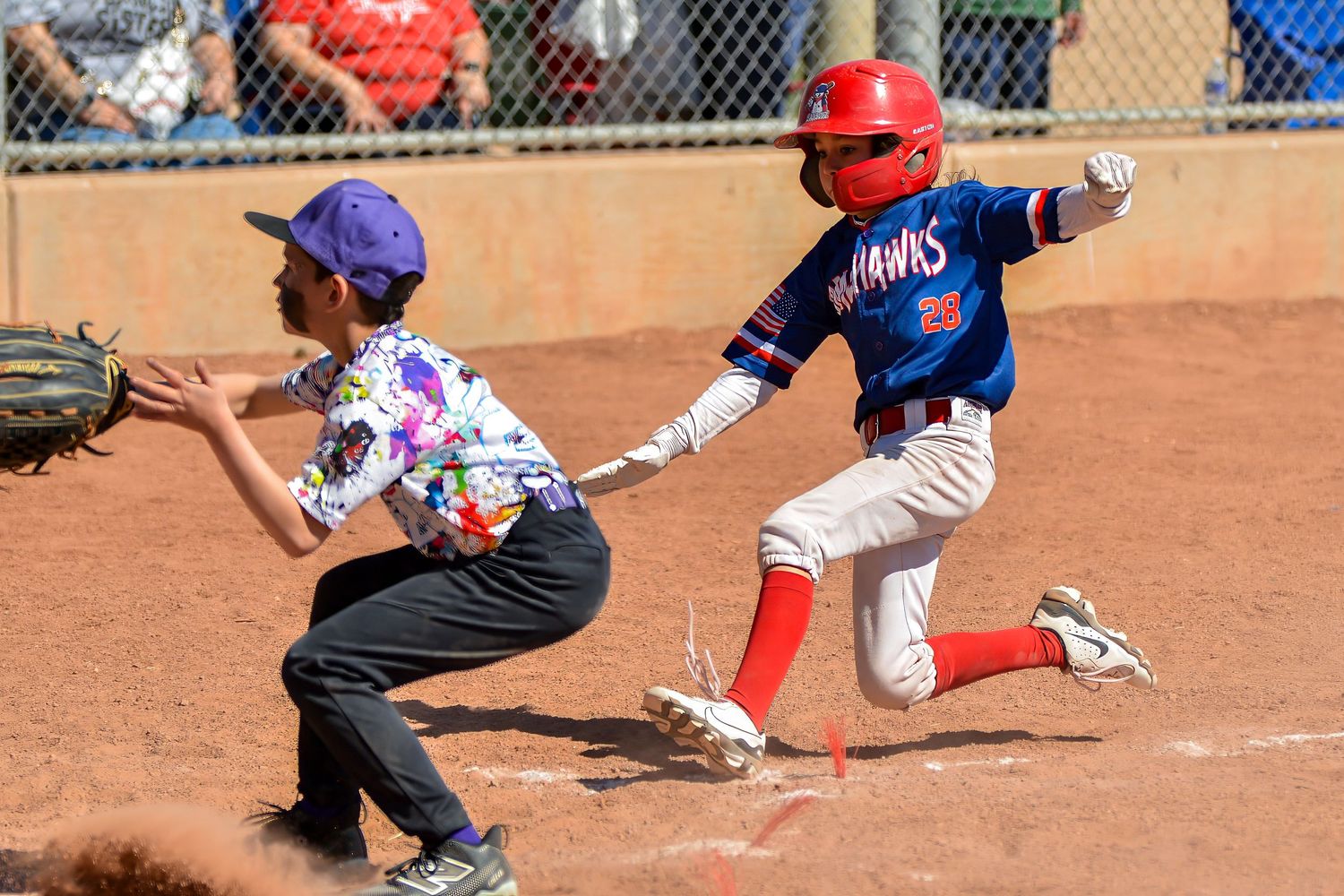 Young baseball player in red helmet slides into base as fielder reaches to make the tag.
