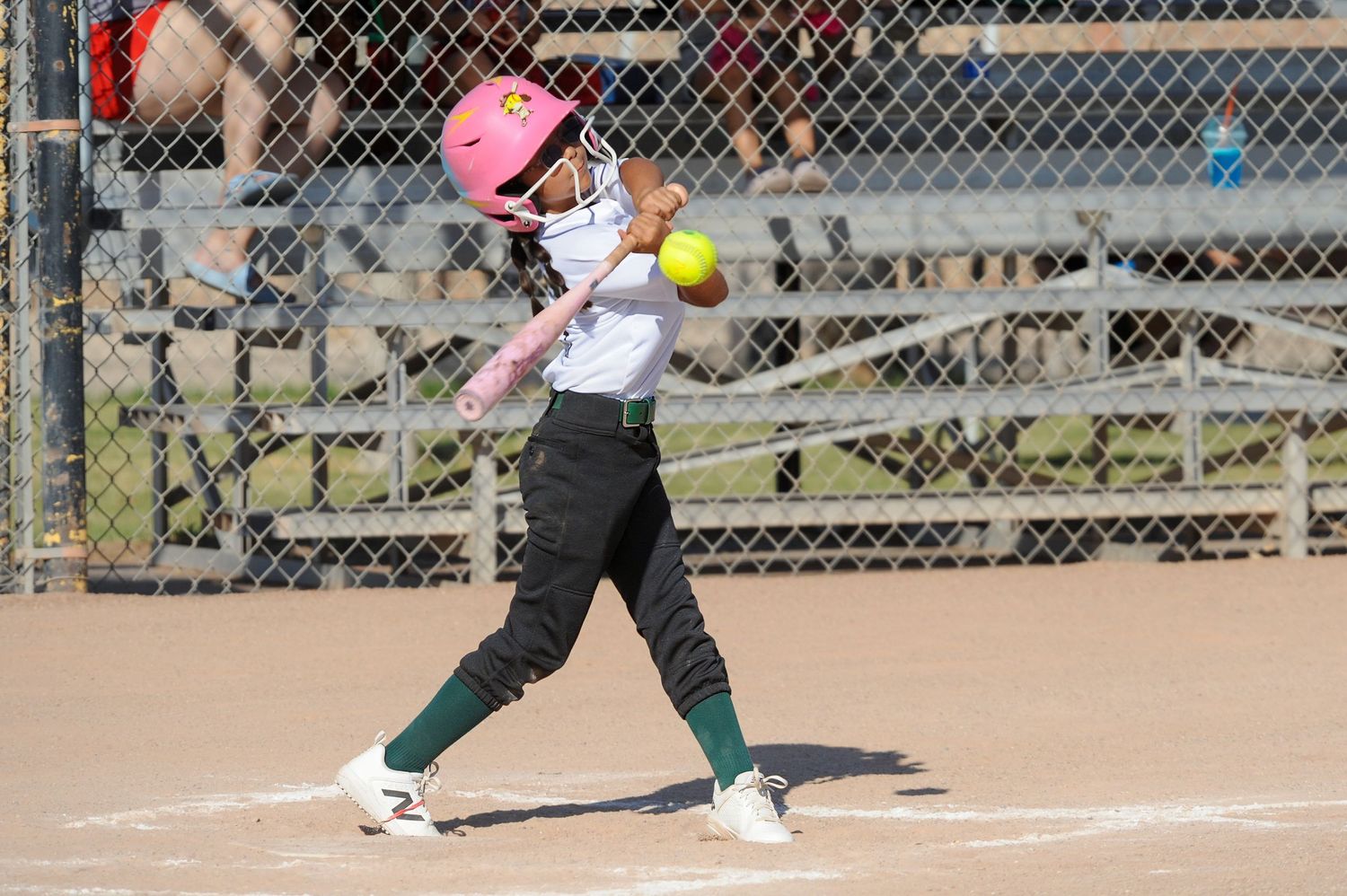 Young softball player in pink helmet swinging bat at yellow ball during a sunny outdoor game.