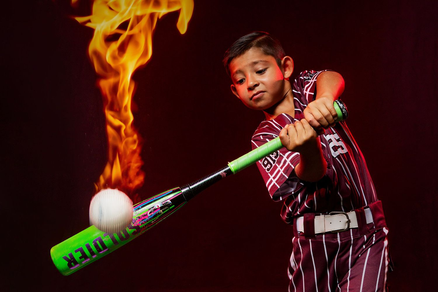 Young baseball player in maroon uniform swinging a green bat at a ball with dramatic fire flames in the background.