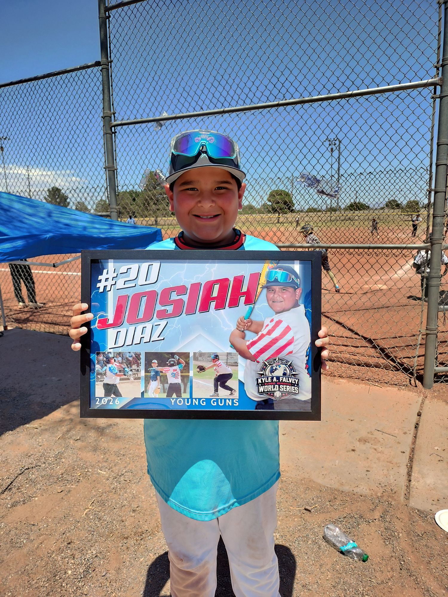 Young baseball player #20 Josiah Diaz proudly holds his framed Young Guns team photo at an outdoor baseball field.