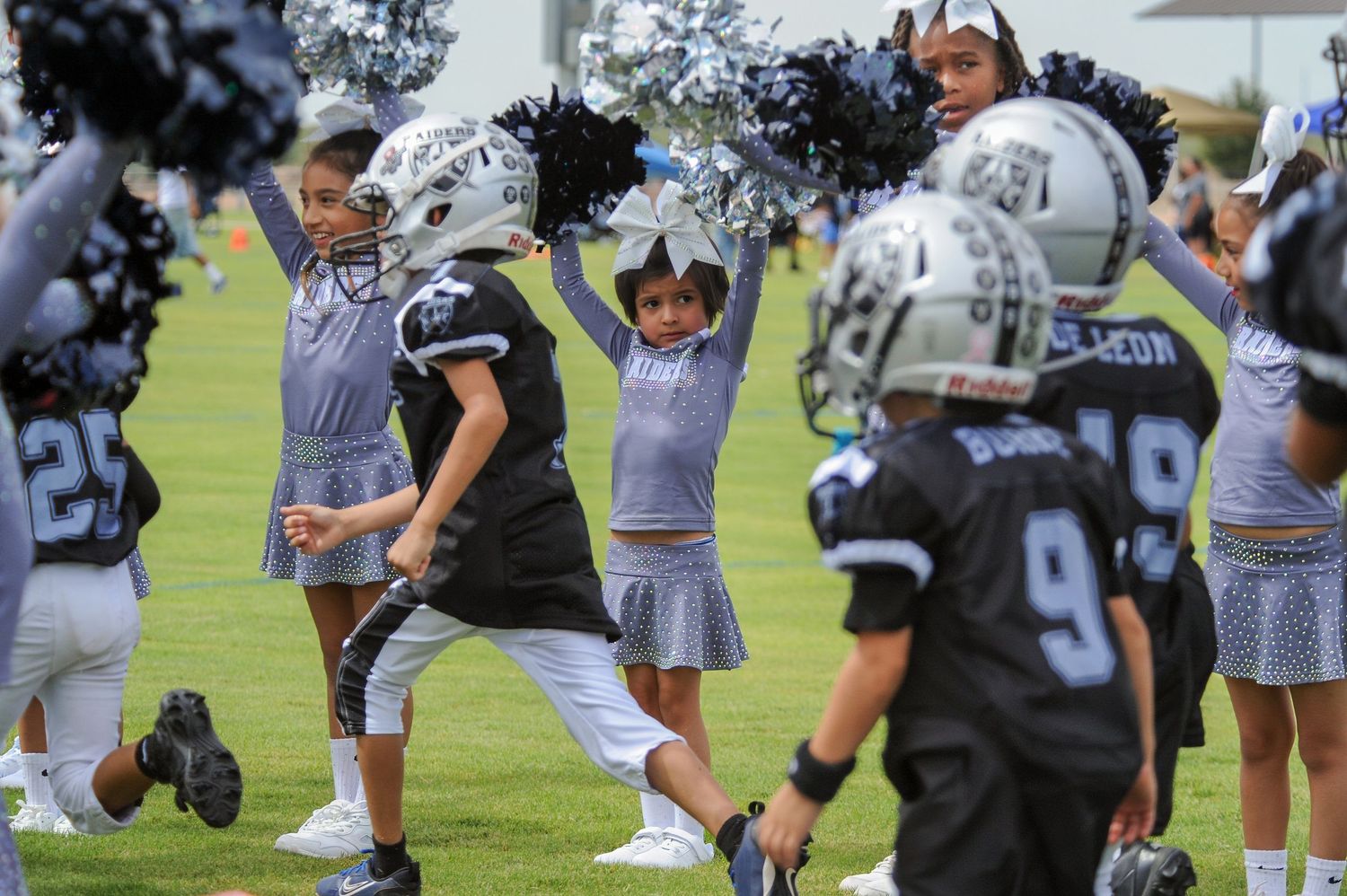 Young cheerleaders in gray uniforms perform with pom-poms alongside youth football players in black helmets on a green field.