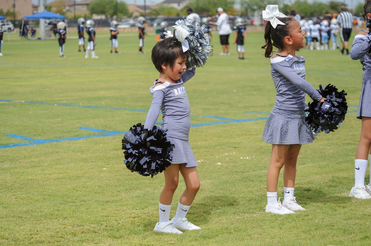 Young cheerleaders in gray uniforms with black pom-poms perform on a green field during a youth football game.