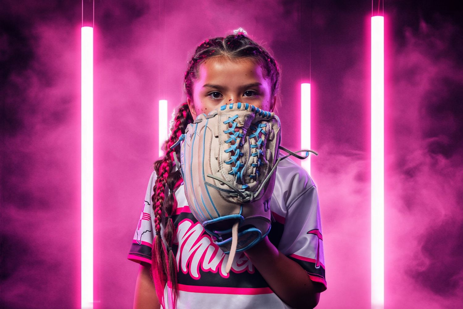 Young softball player in uniform holds glove up, posed against vibrant pink neon lights and smoke backdrop.