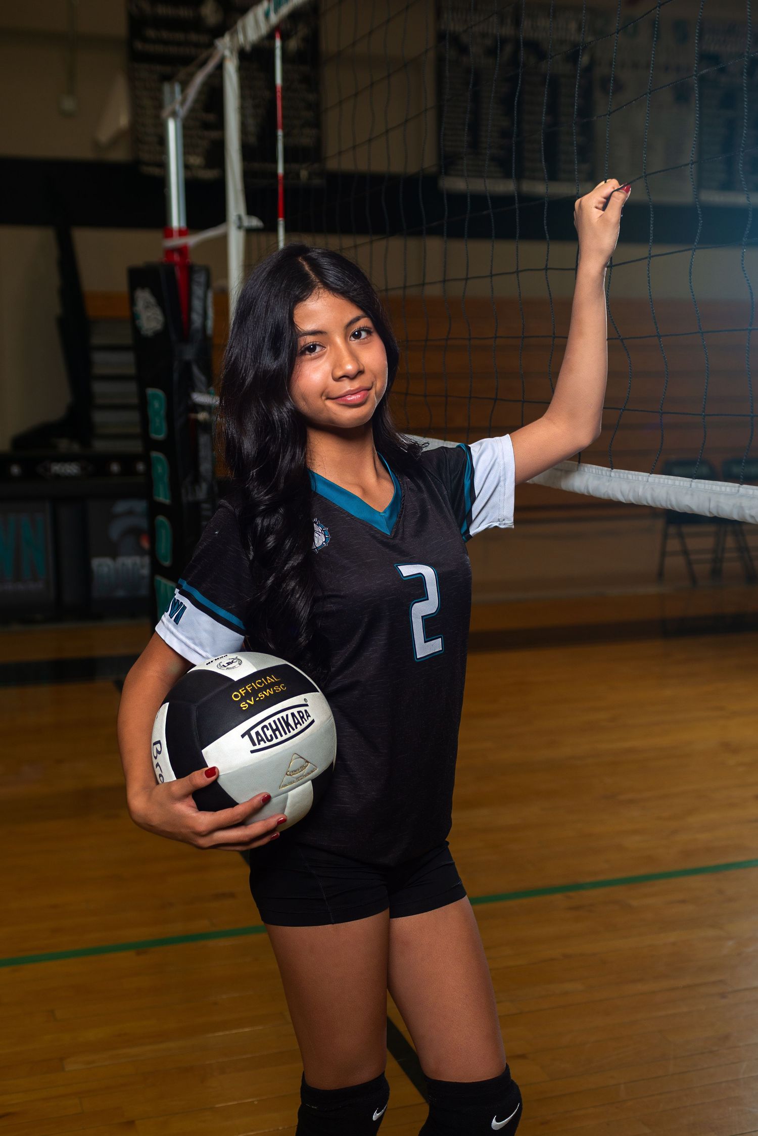 Female volleyball player in black jersey number 2 holds ball beside net in gym.