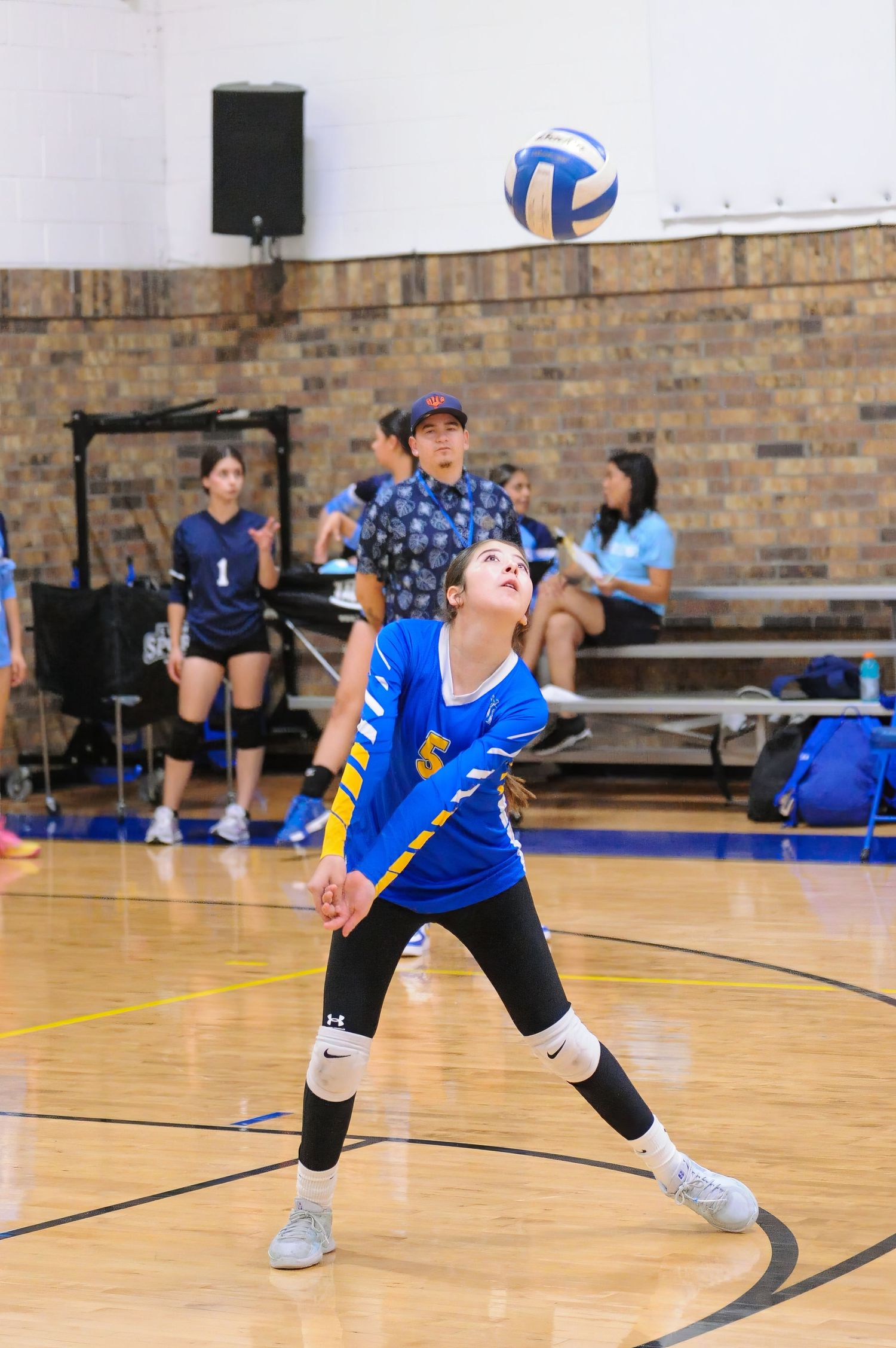 Young female volleyball player in blue uniform preparing to serve the ball during an indoor gym match.