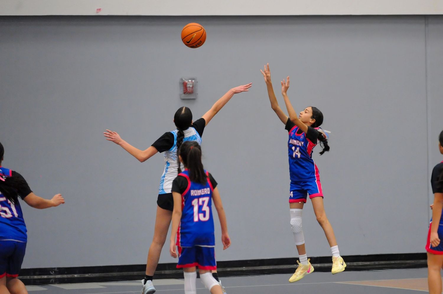 Girls basketball players compete in an indoor game, with one player jumping to shoot the ball over a defender.