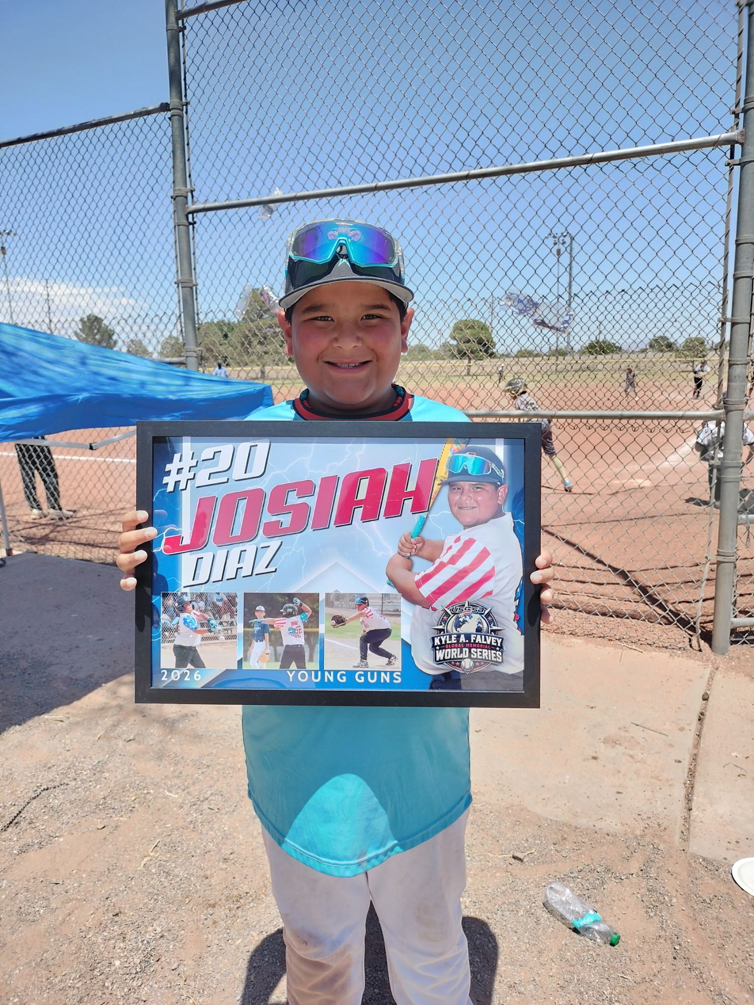 Young baseball player Josiah Diaz, #20, proudly holds a framed Young Guns team photo at an outdoor baseball field.