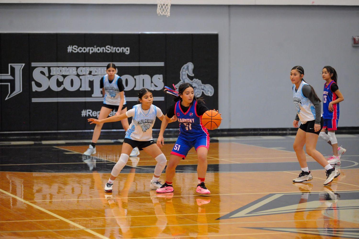 Girls basketball players compete in a gym with Scorpions branding, one dribbling past defenders on a hardwood court.