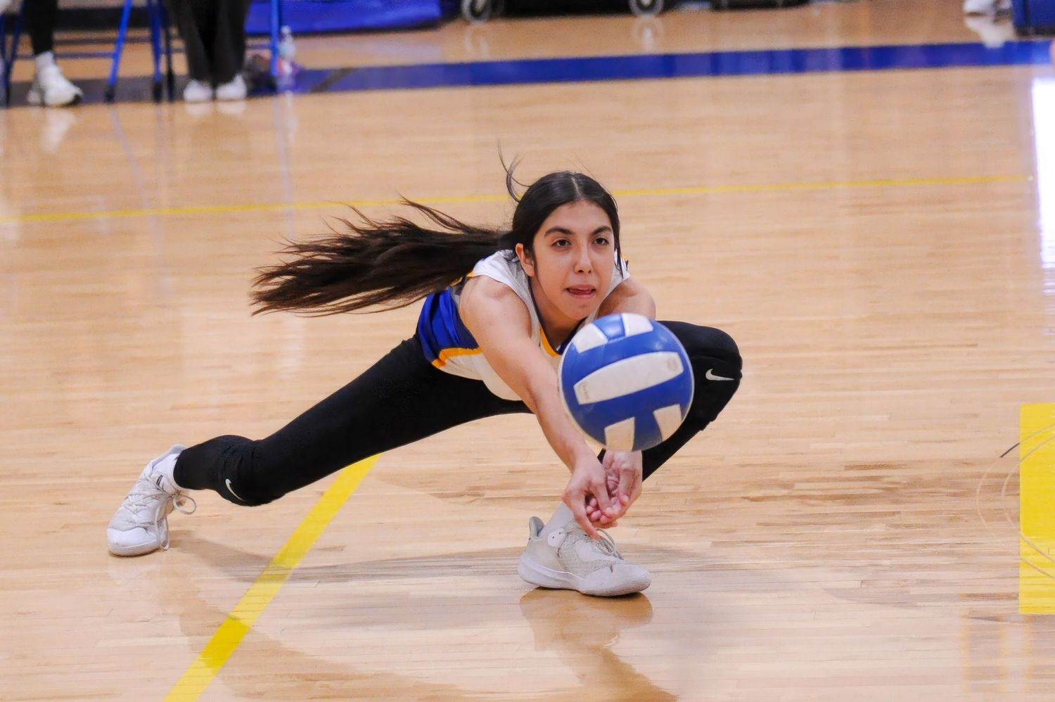 Female volleyball player diving low to dig a ball during an indoor match on a hardwood court.