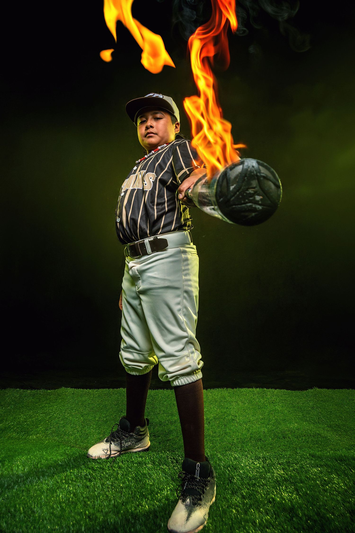 Young baseball player in uniform swinging a flaming bat on green grass with dramatic dark background.