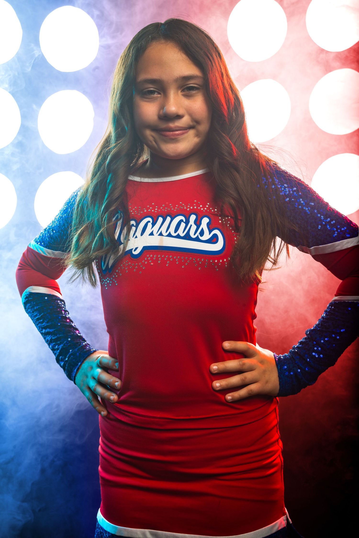 Young girl in a red and blue Jaguars cheerleader uniform poses with hands on hips against a bokeh light background.