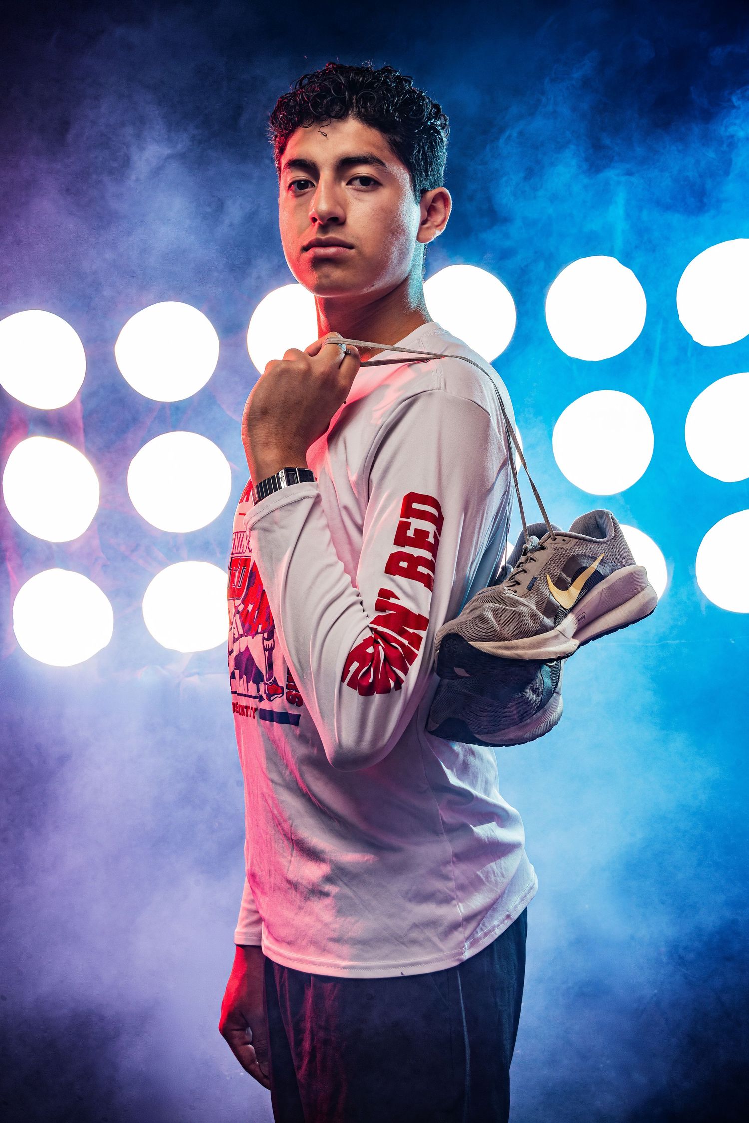 Young athlete in white long-sleeve shirt holds Nike sneakers, posing confidently against dramatic stage lighting backdrop.