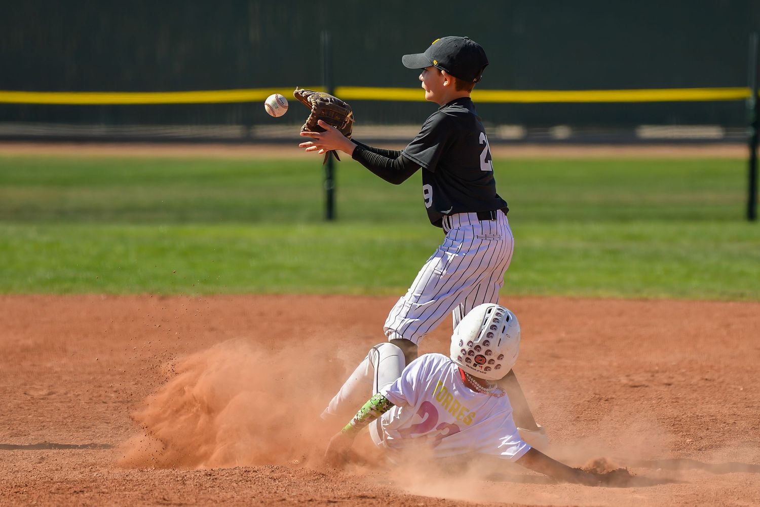 Youth baseball player catches ball at second base as runner slides in during a dusty youth league game.