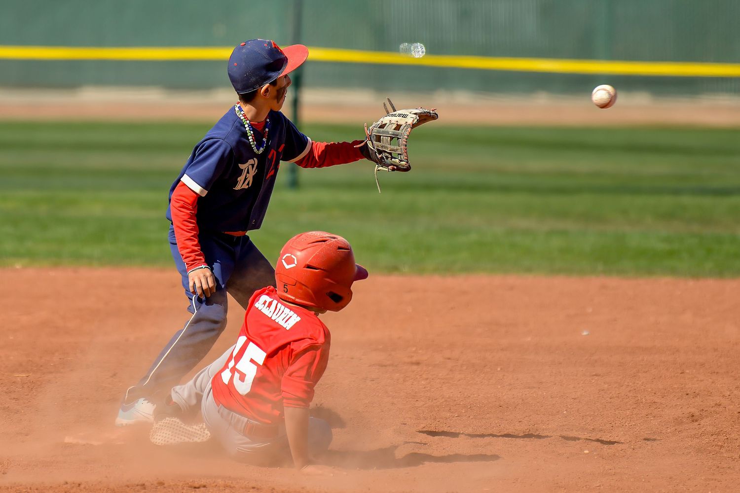 Young baseball player catches a throw as an opposing runner slides into second base in a cloud of dust.