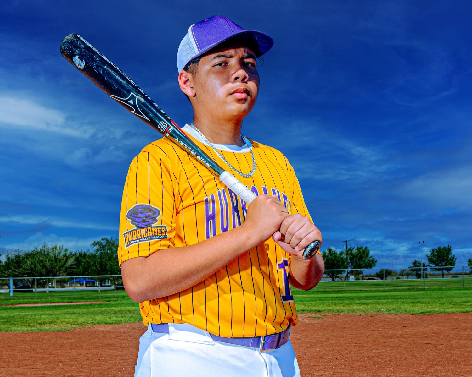 Young baseball player in gold pinstripe Hurricanes jersey and purple cap poses with bat on field.