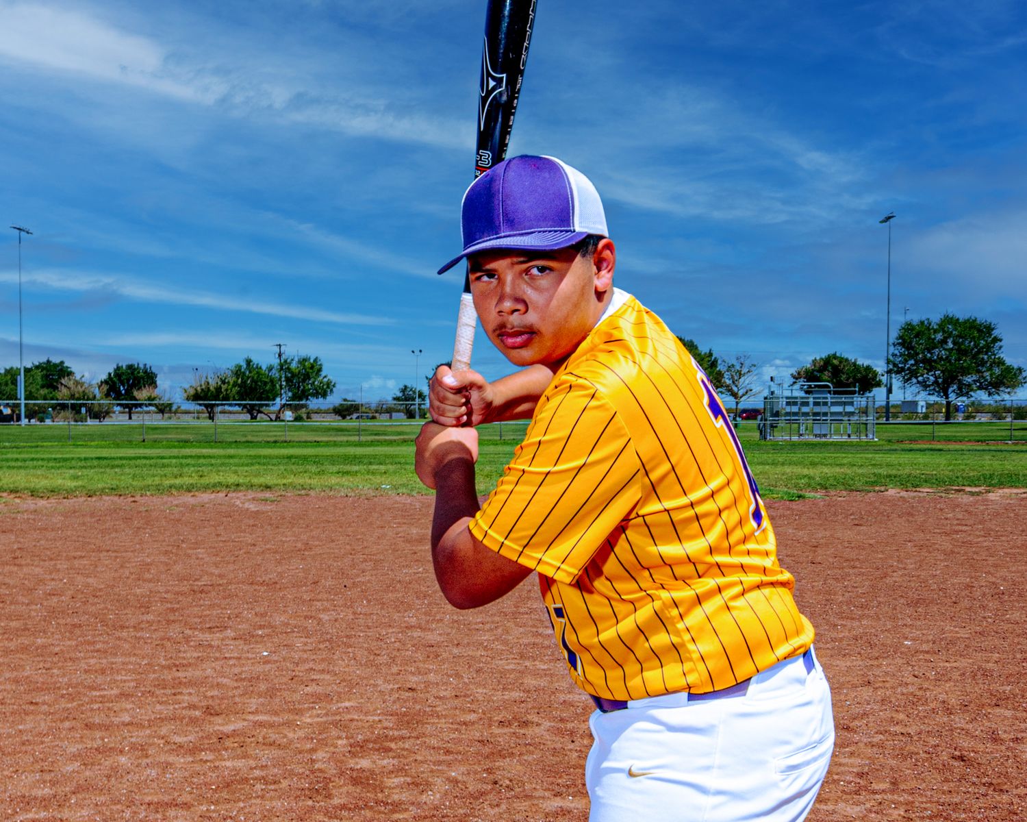 A young baseball player in a yellow pinstripe jersey and purple cap holds a bat in batting stance on a dirt field.