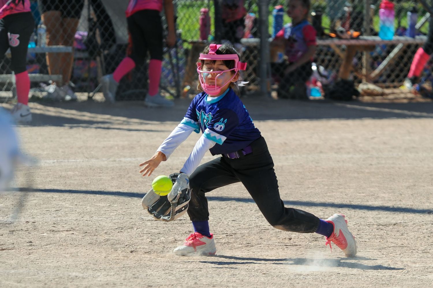 Young softball player in pink helmet lunges to field a ground ball during a youth league game.