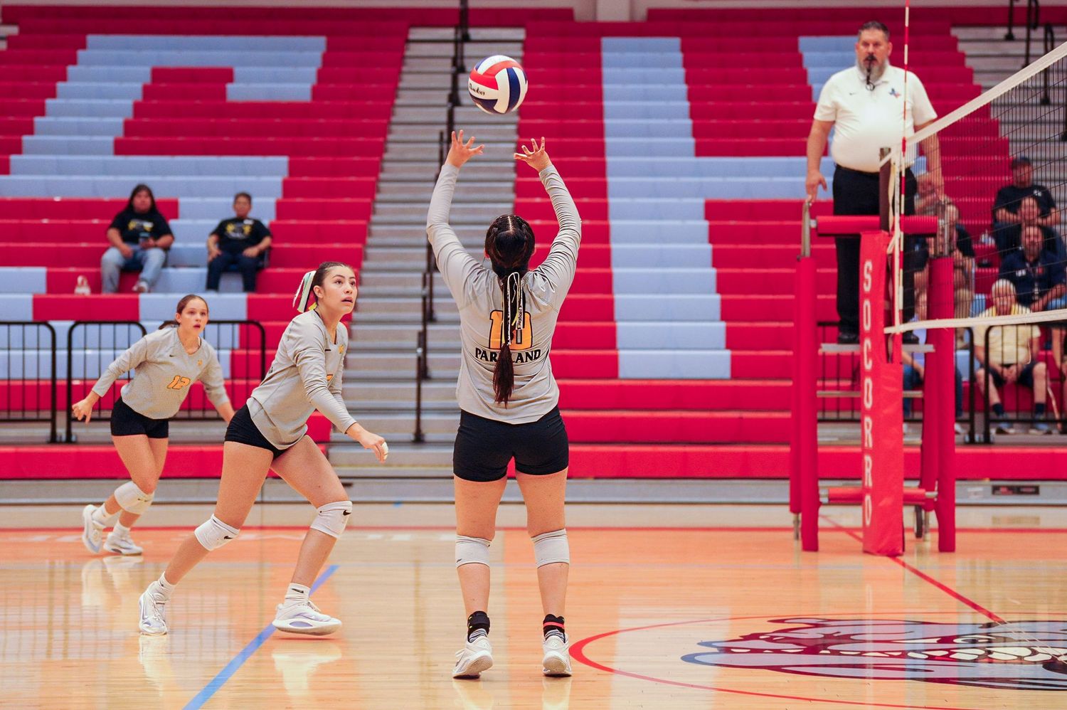 Female volleyball players in gray uniforms compete indoors, with a setter positioning the ball near the net.