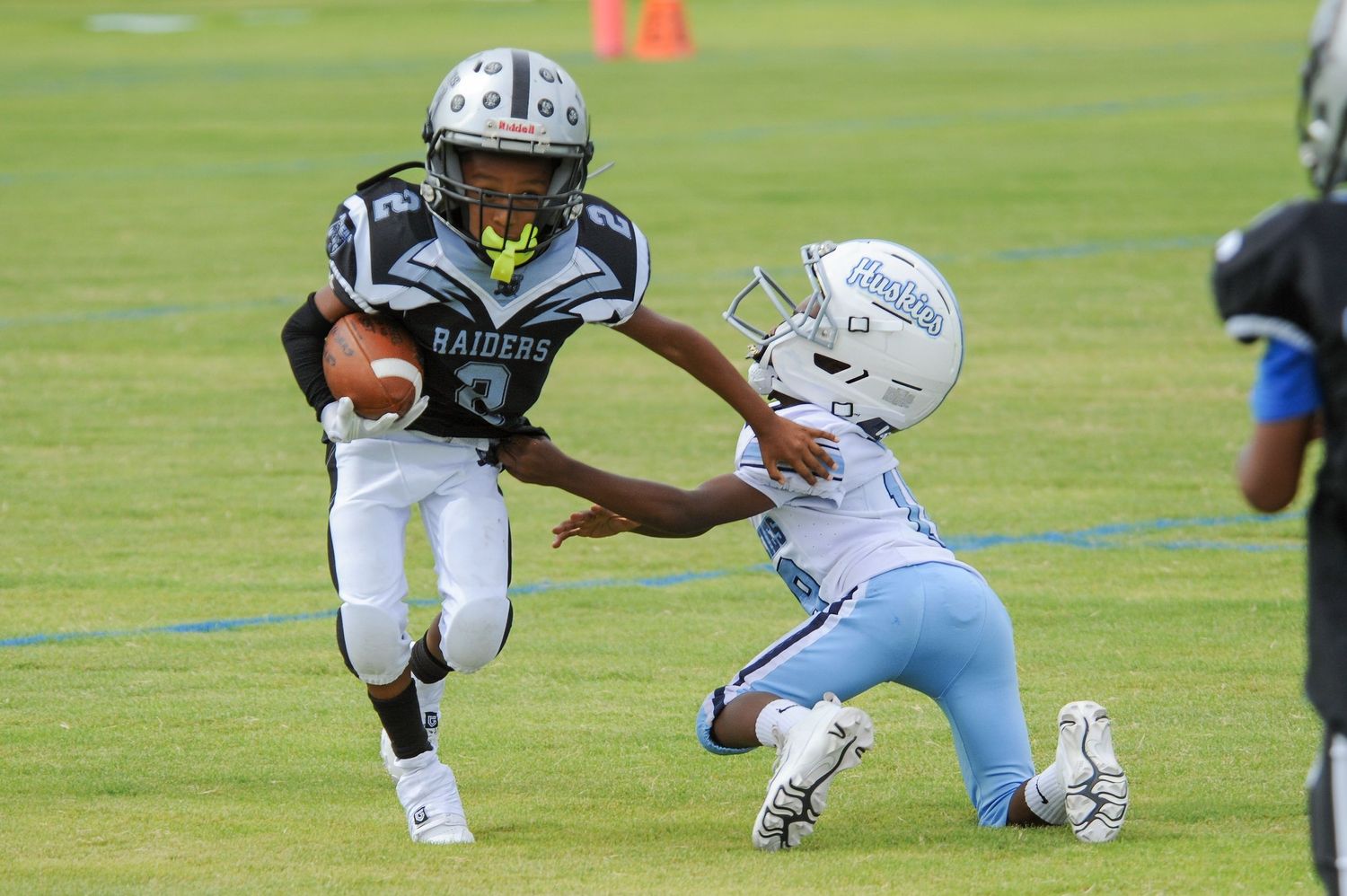 Youth football players compete, Raiders ball carrier evades defender in tackle attempt on green field.