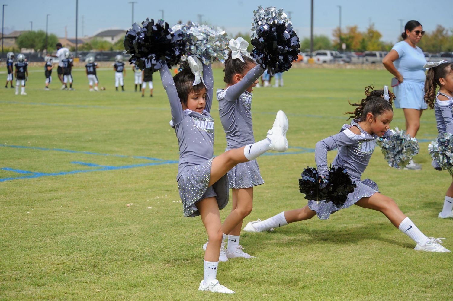 Young cheerleaders in gray uniforms perform energetic routines with silver pom-poms on a green field.