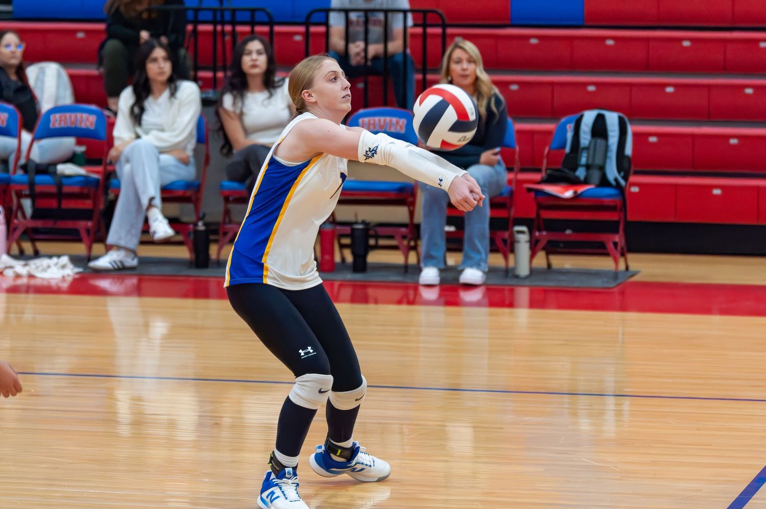 Volleyball player in blue and gold uniform performs a dig during an indoor match on a red court.
