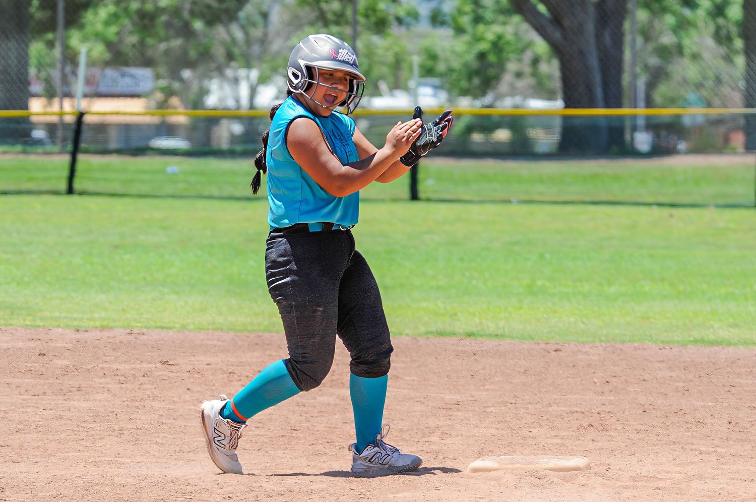 Softball player in teal uniform celebrates at second base during a sunny outdoor youth league game.