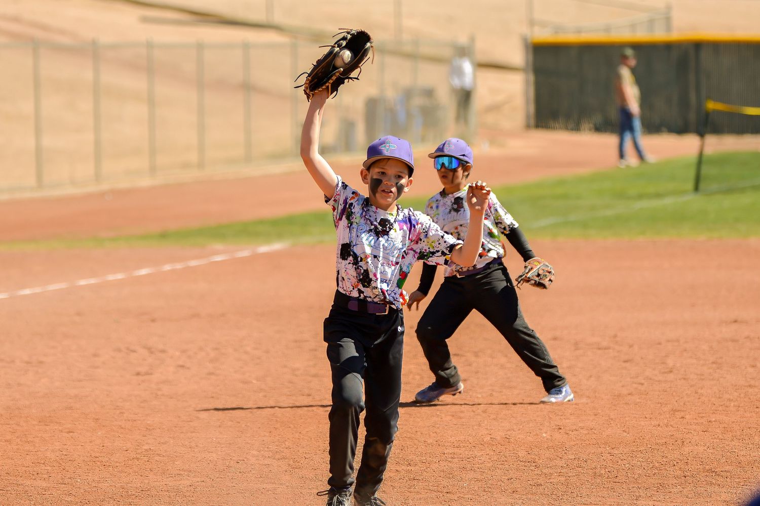 Two youth baseball players celebrate on the field, one raising a glove in victory during a game.