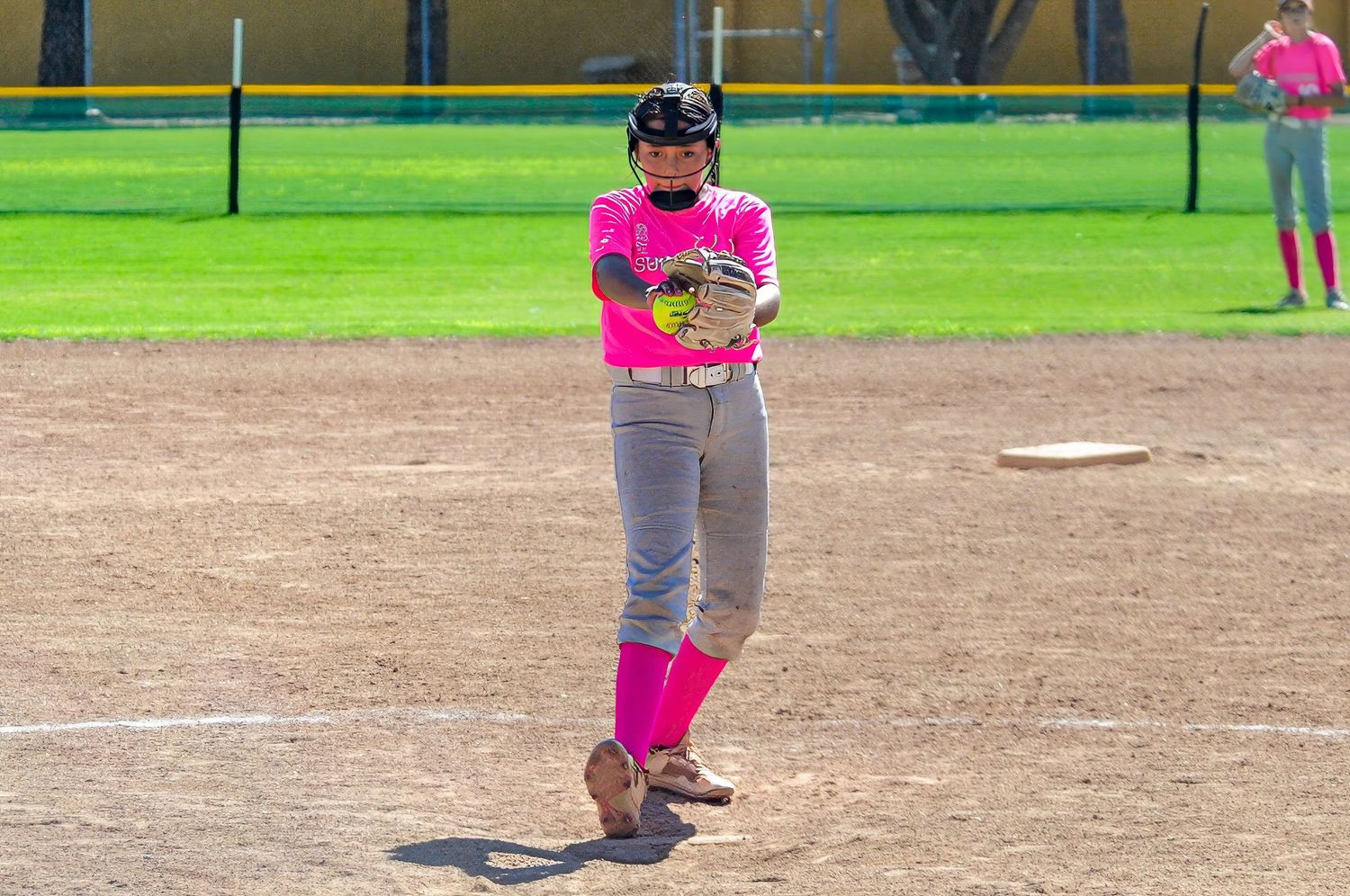 Female softball player in pink uniform and pink socks standing ready on the pitcher's mound outdoors.