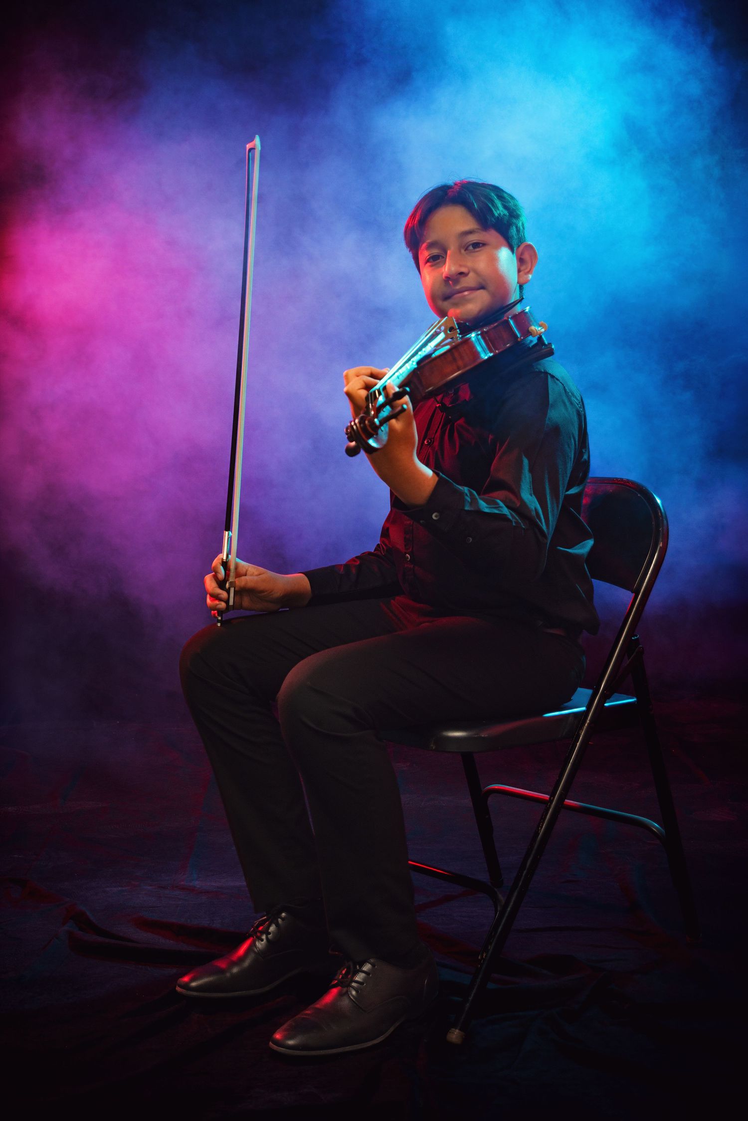 Young violinist sitting on chair holding bow, performing under dramatic pink and blue smoky stage lighting.