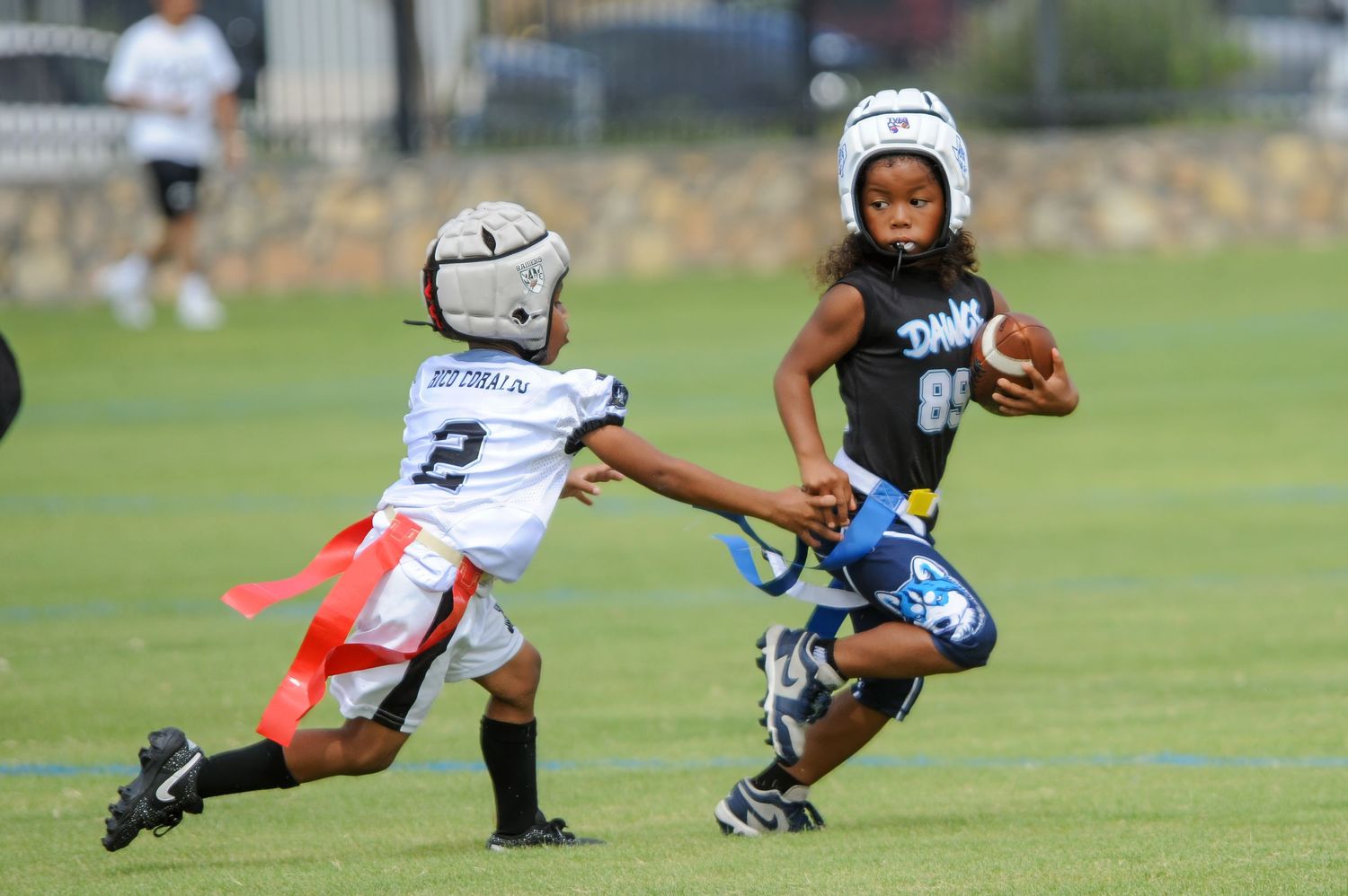 Two young boys playing youth flag football game on green field, one carrying ball while other attempts to grab flag.