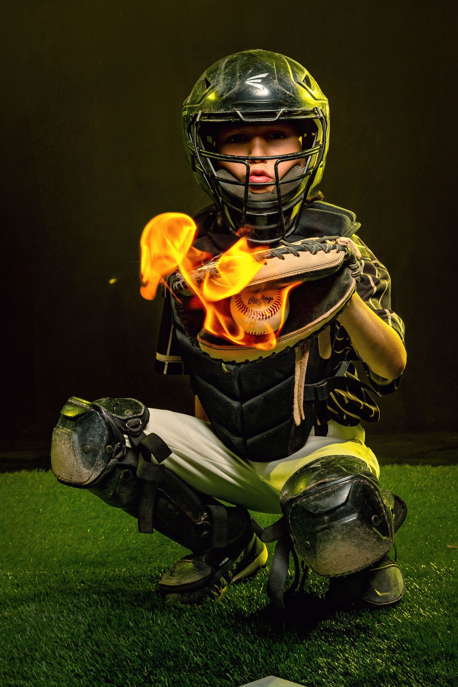 Young baseball catcher in full gear crouching with a flaming ball in glove on dark background.