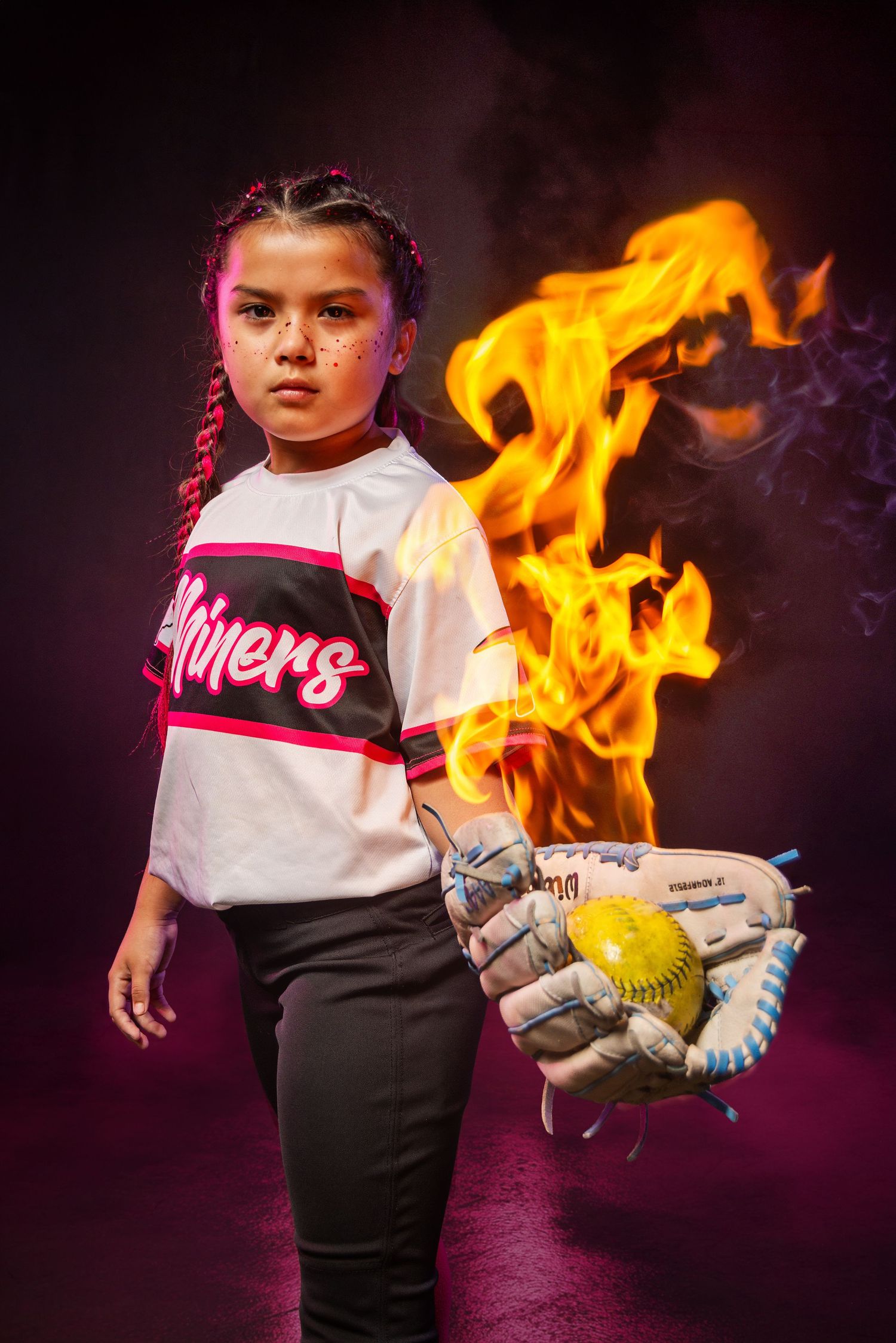 Young girl in Miners softball jersey holds glove with yellow ball, dramatic flames behind her on dark background.