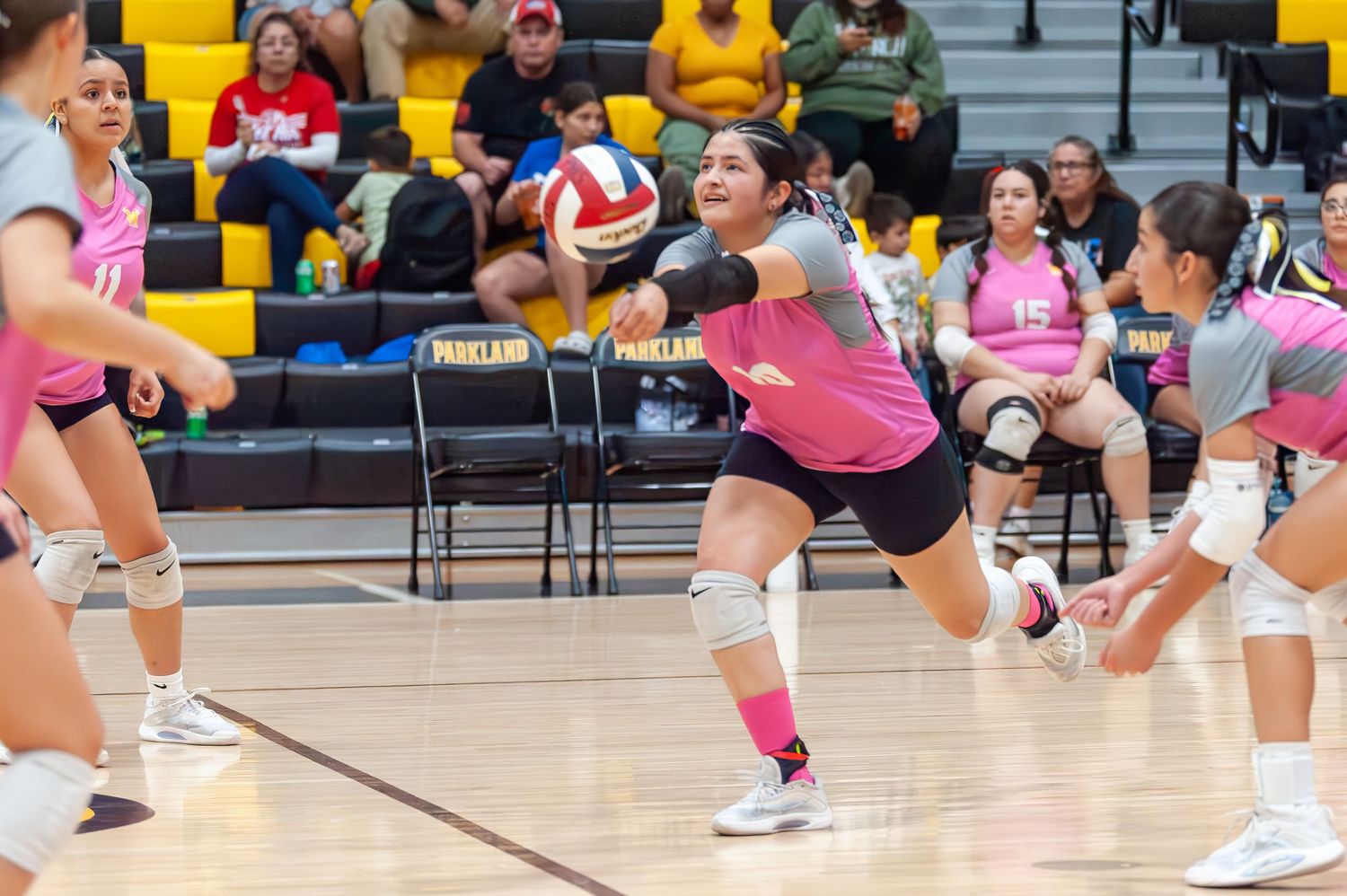 Female volleyball player in pink uniform dives to dig ball during indoor high school volleyball game.
