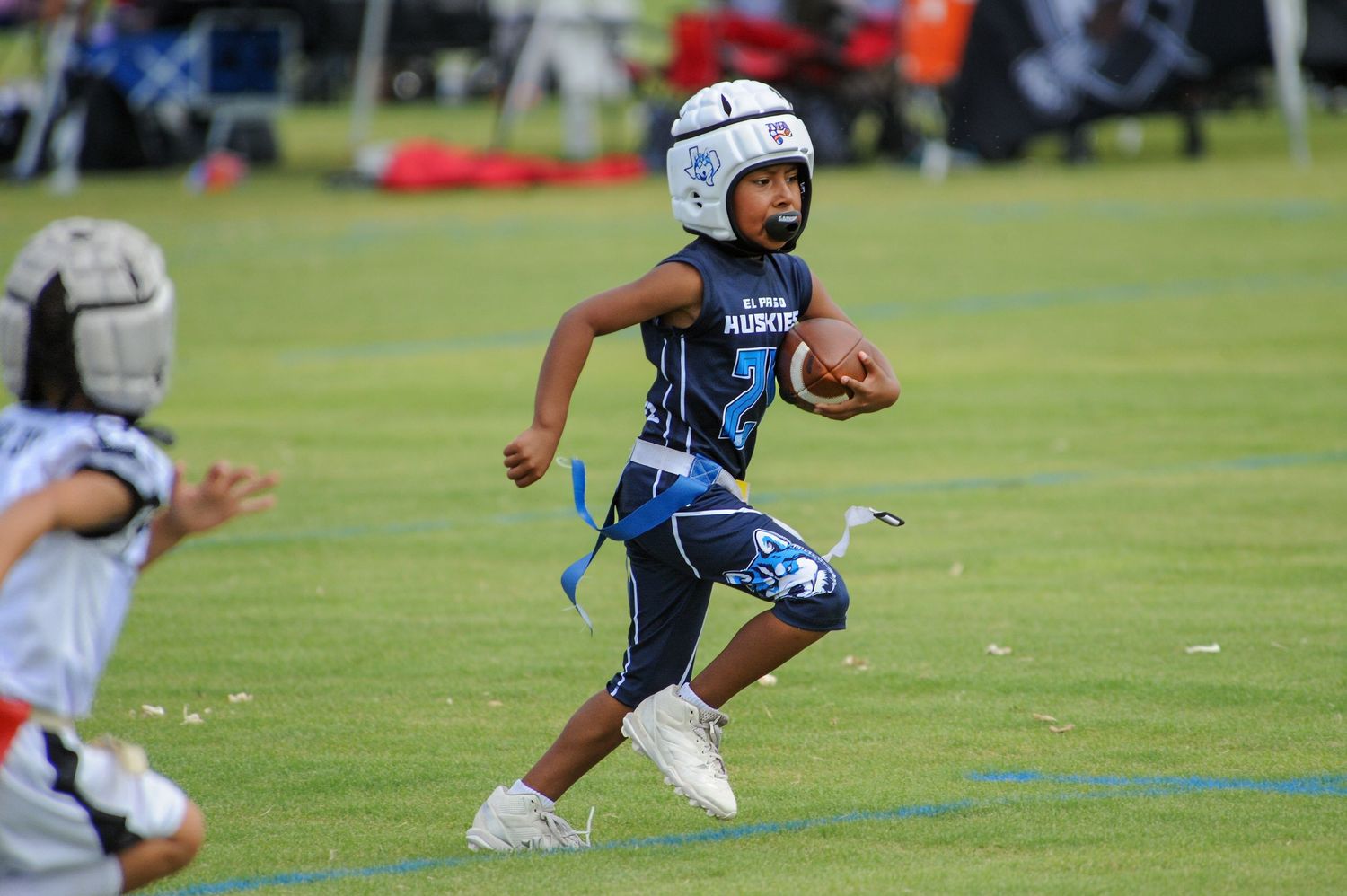 Young child in blue flag football uniform running with the ball during a youth league game on a green field.