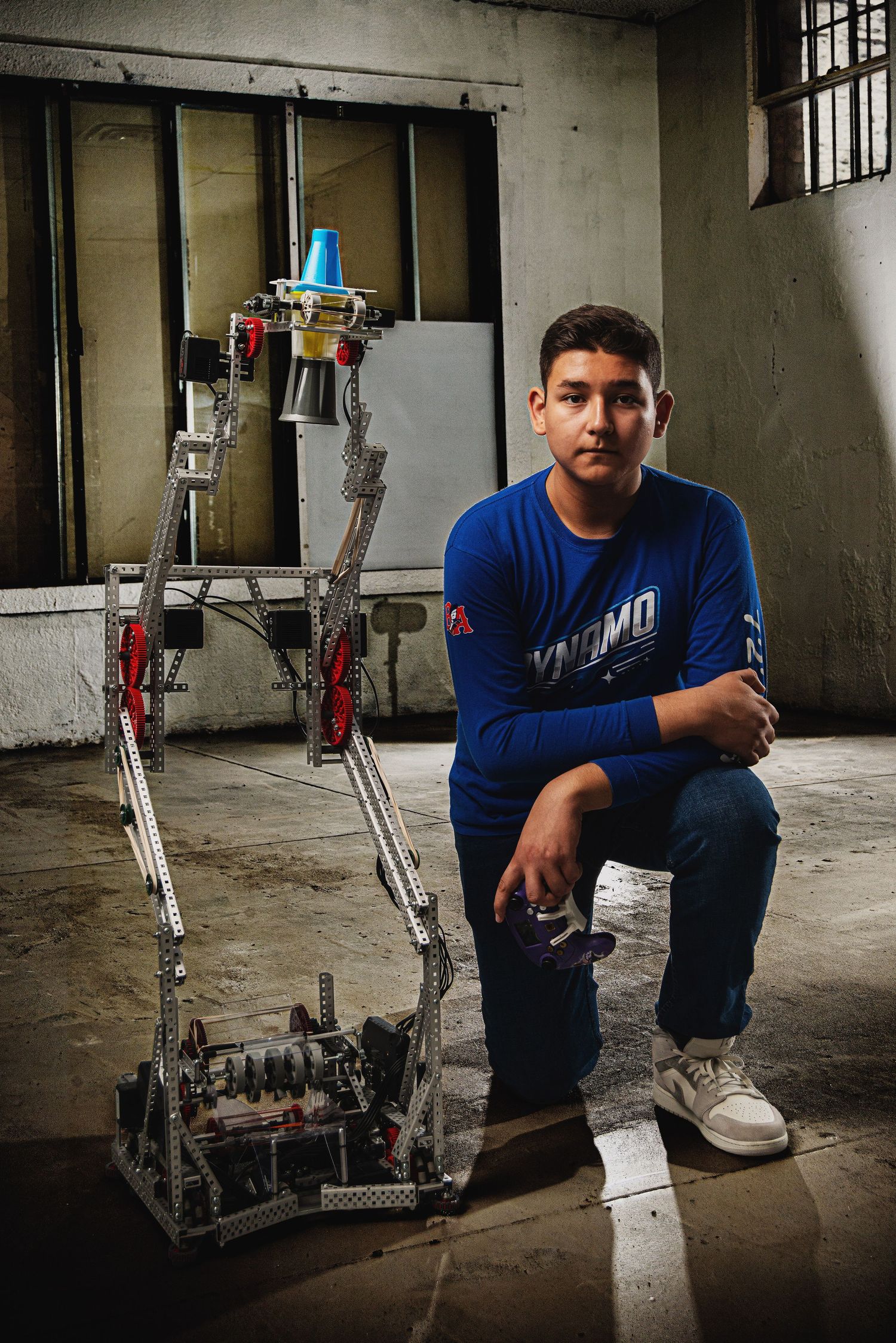A young student in a blue shirt poses with a tall metal VEX robotics competition robot in an industrial setting.